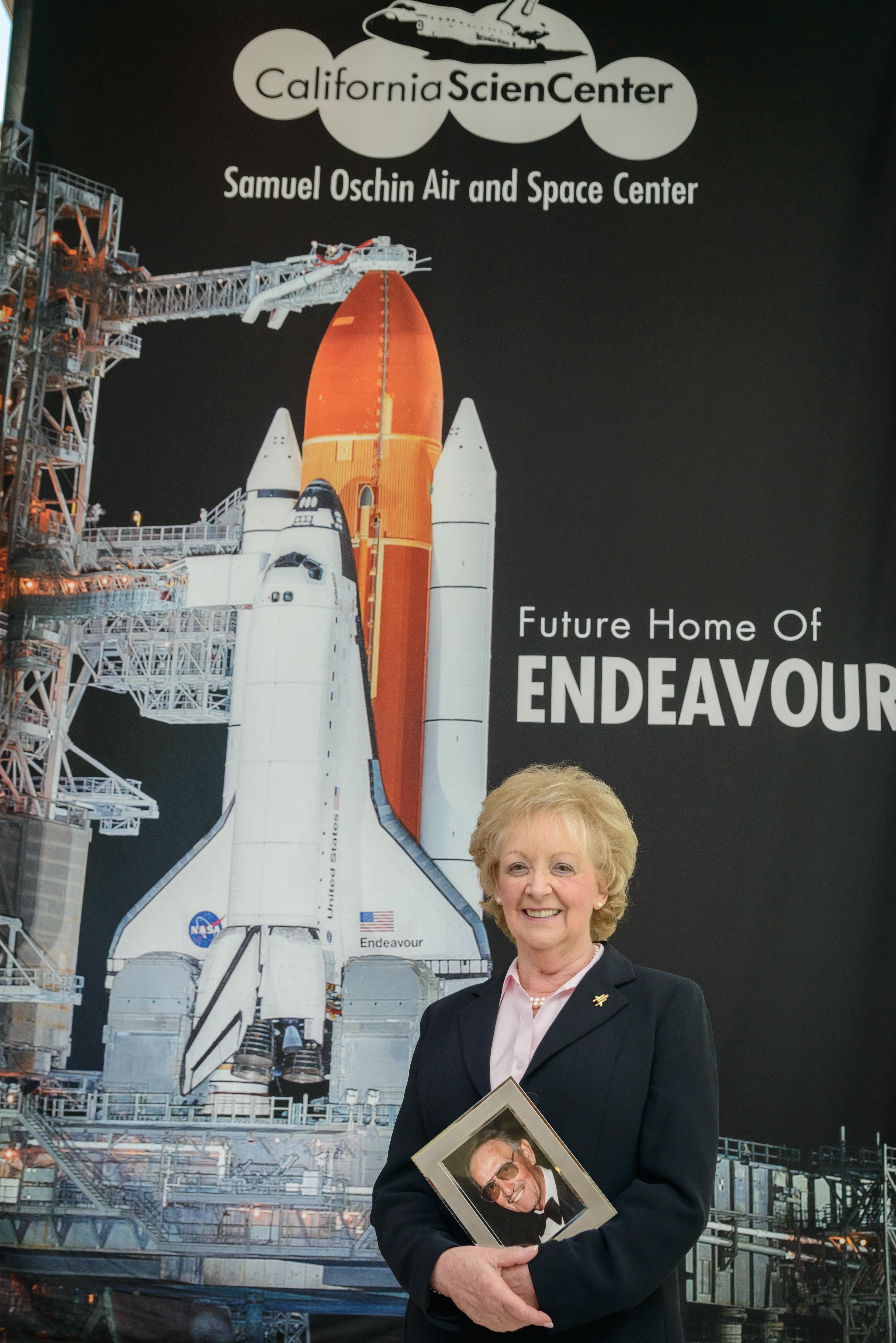 Lynda Oschin standing in front of large banner banner that says "Future home of Endeavour" while holding a portrait of Samuel Oschin