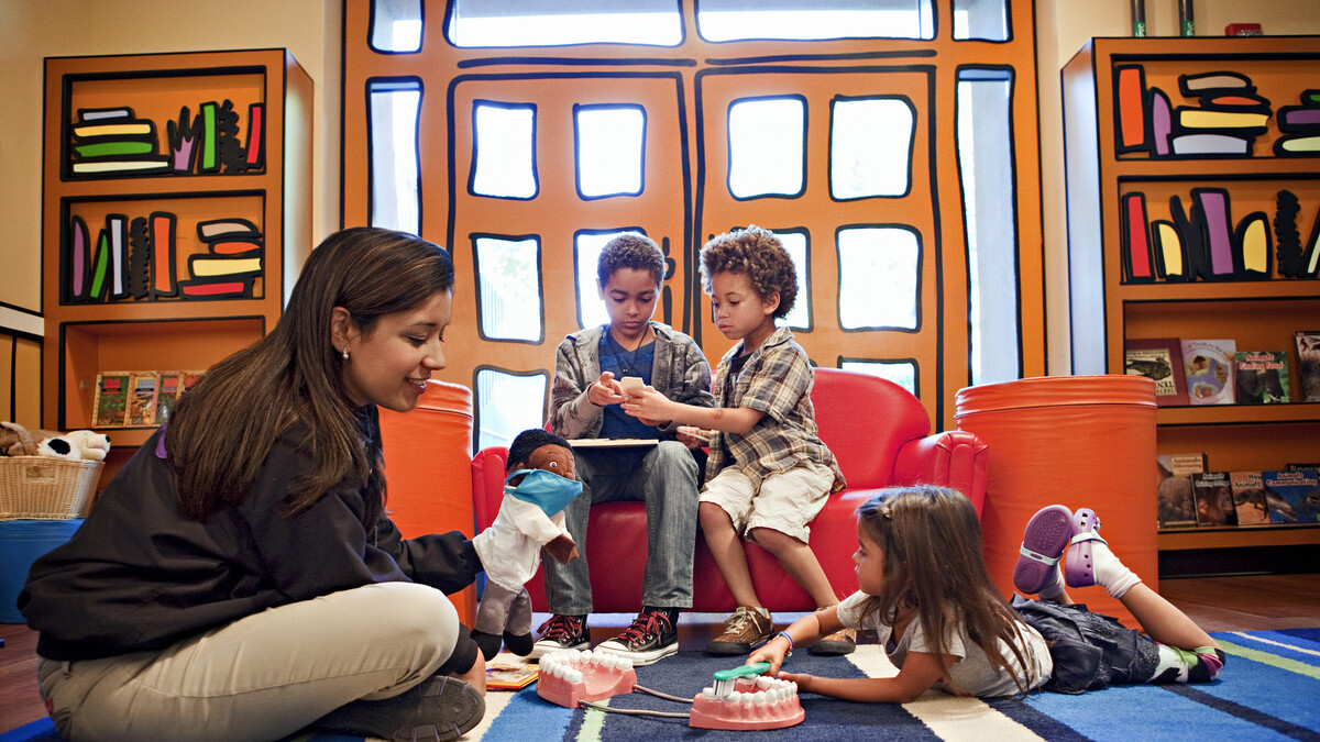 Guest Services facilitator sits on floor with puppet in hand and plays with young children in the Ecosystem Discovery Room reading area