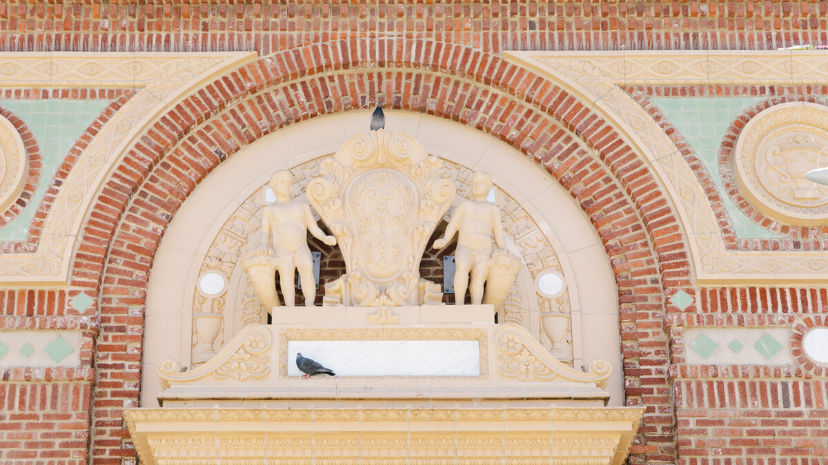 Two pigeons sit atop an intricate concrete architectural decor over the North Patio entrance door. Architectural decor features two figures and a flourish surrounded by arched brick.