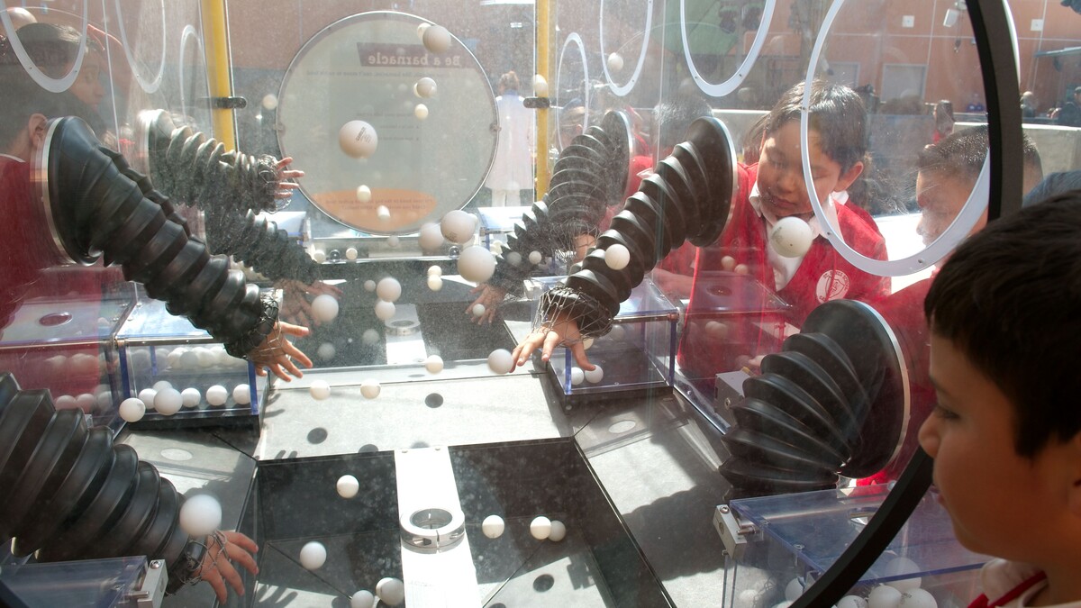 People stick their hands into bendable tubes to reach in a large acrylic box to grab ping-pong balls