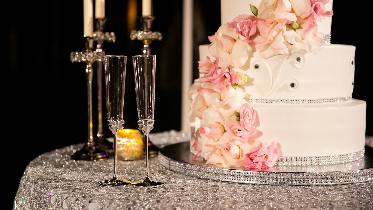 Four tiered wedding cake decorated in pink flowers sits atop a silver sequined cocktail table
