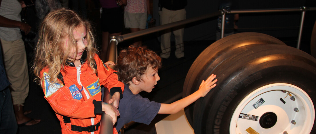 A girl in an astronaut costume with a boy touching space shuttle tires