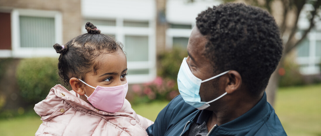 A masked parent holds their kid, who is also wearing a mask